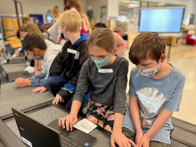 Students collaborating on laptops during a classroom activity, wearing masks for safety with a SMART Board in the background.