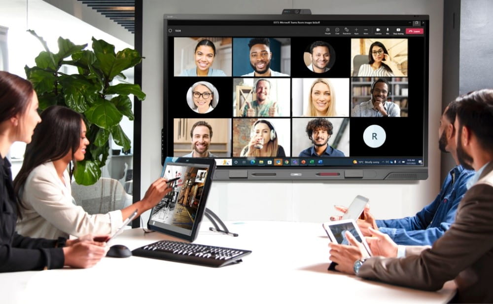 Hybrid meeting in progress with in-person team members interacting with remote colleagues through a video conferencing platform on a SMART interactive display.