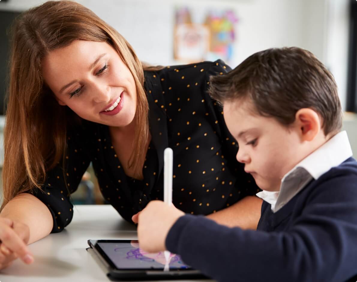 Teacher guiding young student using a digital tablet in classroom.